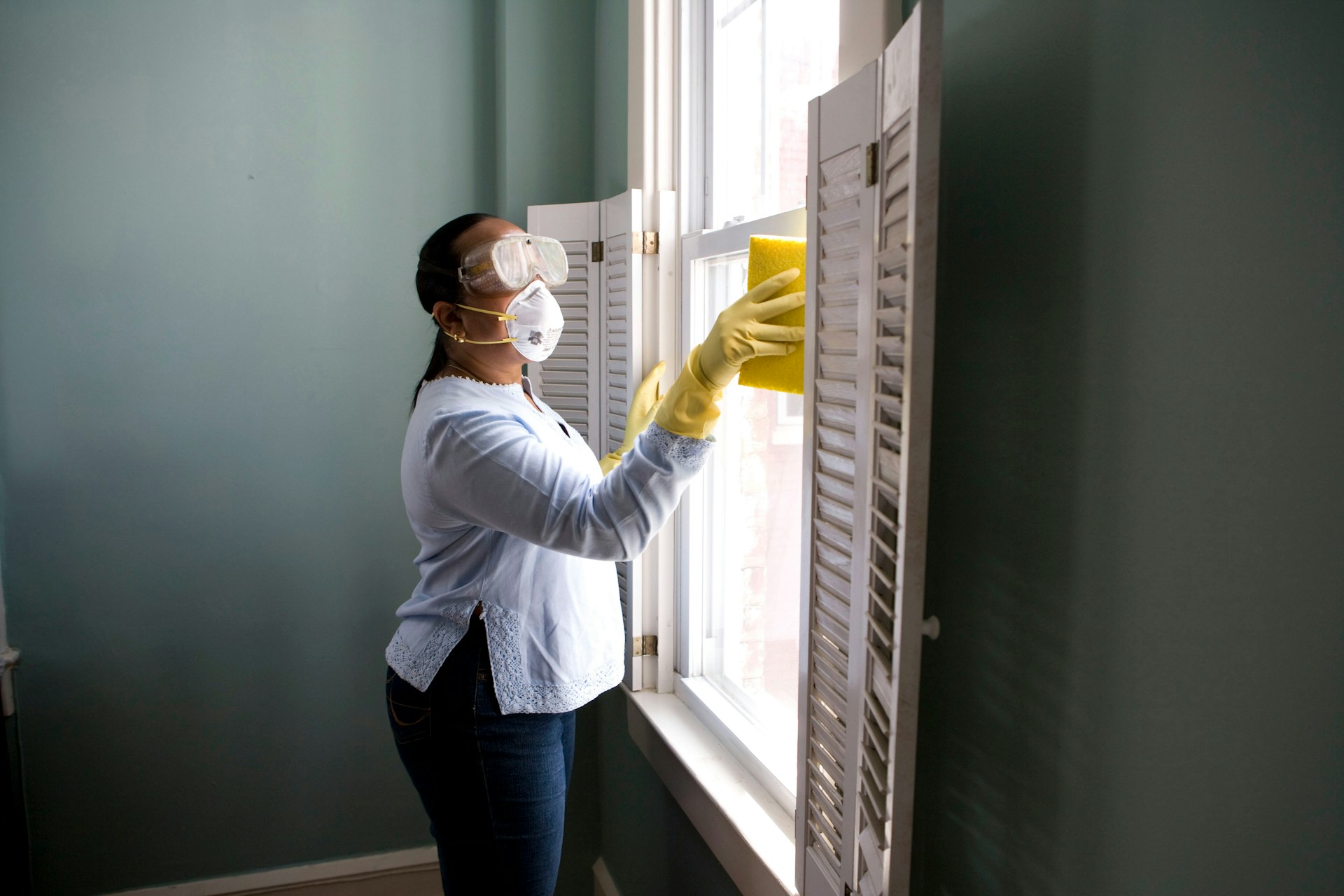 A woman in a white shirt sprays and wipes a window in a bright home interior